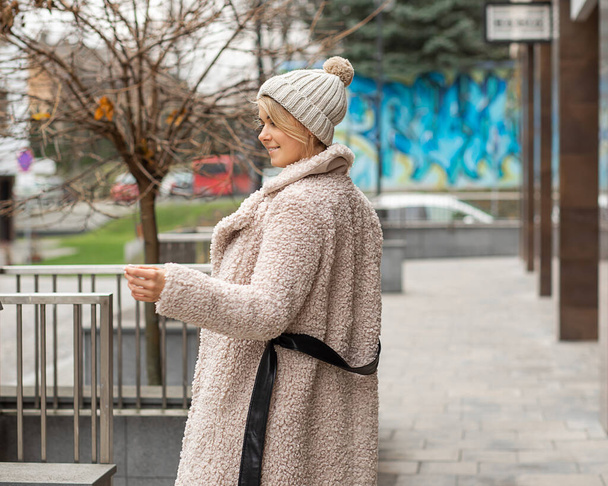 Young cute blonde woman in beige fur coat is happy in city, cheerful smiling to side, rear view, knitted hat. Lifestyle photo, street style - Valokuva, kuva