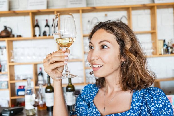 Χαμογελαστή γυναίκα δοκιμάζει το νεαρό λευκό κρασί σε ένα οινοποιείο κατά τη διάρκεια μιας ειδικής περιήγησης. Η έννοια της ανθοδέσμες και νέες γεύσεις από τις καλύτερες ποικιλίες σταφυλιών και επιτραπέζιων ποικιλιών. - Φωτογραφία, εικόνα