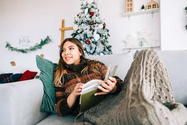 Young caucasian woman relaxing on a sofa at home and reading book in Christmas holidays. - Zdjęcie, obraz