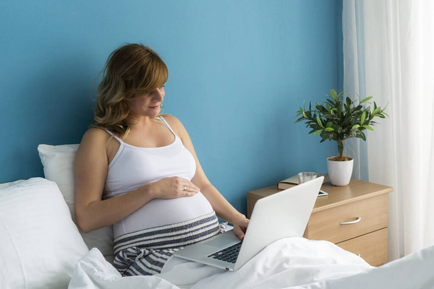 Een glimlachende en gelukkige zwangere blanke vrouw in een zittende positie in haar bed terwijl ze aan haar laptop werkt. Eén hand zit op haar buik.. - Foto, afbeelding