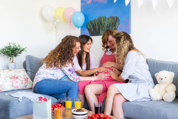 Egy baráti társaság a nappaliban ünnepel a barátjával, aki gyereket vár. Mindenki boldog és mosolyog, mind a négyen a hasán tartják a kezüket a babával.. - Fotó, kép