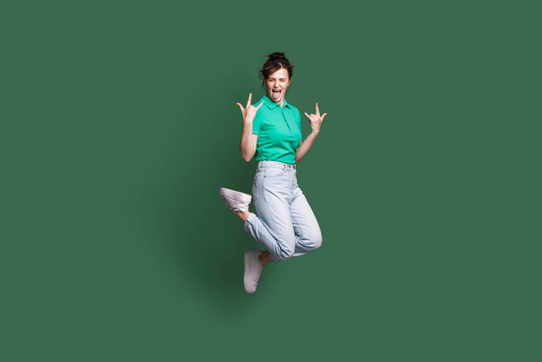Full size photo of young crazy caucasian woman jumping showing tongue. Rocknroll sign. Isolated on green color background - Фото, зображення