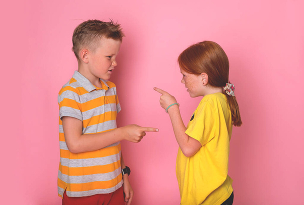 2つの怒っている子供たちの弟と妹の顔に直面して立って、お互いに指を指して叫んで非難する。ピンクの壁に対して学校の子供。女の子と男の子の口論 - 写真・画像
