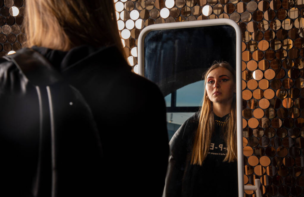 A teenage girl with long blonde hair, in black clothes and with bright makeup looks at herself in the mirror. - Photo, image