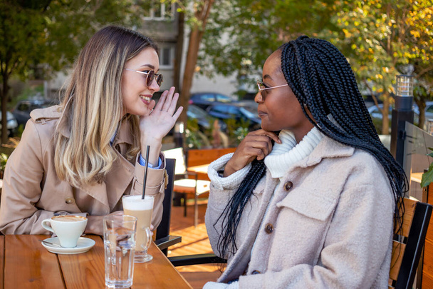 Kaksi nuorta tyttöystävää nauttivat kahvista yhdessä, istuvat pöydässä, juttelevat, kuiskailevat ja juoruavat. Monimuotoisuus ja vapaa-ajan käsite. - Valokuva, kuva