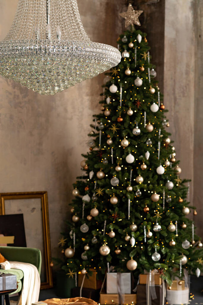 Fondo de año nuevo. Habitación loft de Navidad interior. Iluminación, una gran lámpara de araña de cristal, velas e iluminación interior caliente, guirnaldas cerca del árbol de Navidad con regalos. Enfoque selectivo suave. - Foto, Imagen