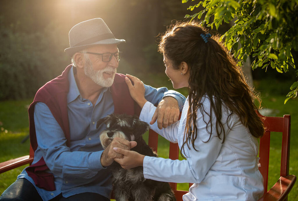 Homme âgé avec chien assis dans le parc tandis que jolie infirmière en manteau blanc tenant la main sur son épaule, prenant soin du patient - Photo, image