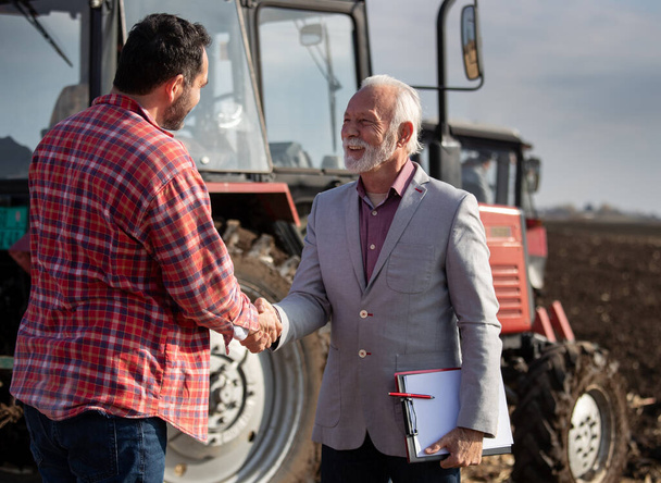 Alto rappresentante di vendita e agricoltore maturo che stringe la mano sul campo davanti al trattore - Foto, immagini