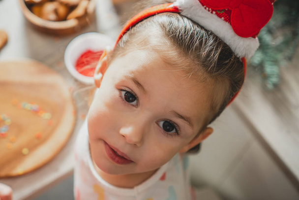 Klein donkerharig meisje van 3 jaar oud in Santa haarband kijkt op naar camera in de keuken. Vrolijk Kerstfeest, Gelukkig Nieuwjaar - Foto, afbeelding
