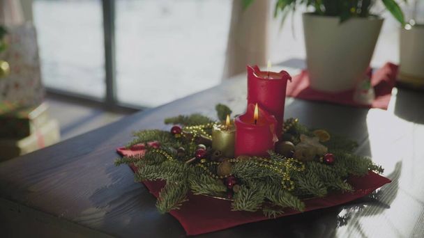 2本の赤いキャンドルと1本の黄金のキャンドルを持つ常緑の花輪。背景にはクリスマスツリーが飾られています。クリスマスの伝統と休日の前夜のキリスト教徒の習慣. - 写真・画像