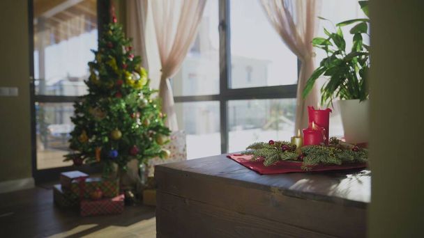 Una corona de hoja perenne con dos velas rojas y una vela dorada. En el fondo, un árbol de Navidad decorado. Las tradiciones y costumbres navideñas de los cristianos en vísperas de la fiesta. - Foto, Imagen