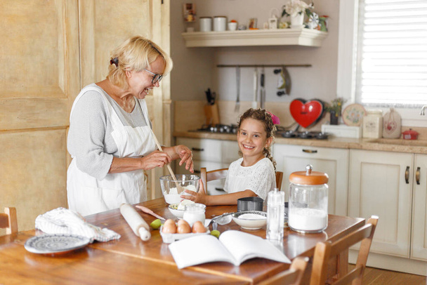 Γιαγιά και όμορφη αξιολάτρευτη εγγονή προετοιμάσει μαζί στην κουζίνα, με τα συστατικά στο τραπέζι. - Φωτογραφία, εικόνα