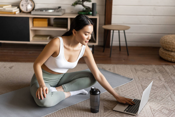 Spor ve yaşam tarzı konsepti. Gülümseyen genç Asyalı kadın evde yoga yapıyor, laptopta meditasyon kursunu takip ediyor, zen pozunda yer paspasında oturuyor. - Fotoğraf, Görsel