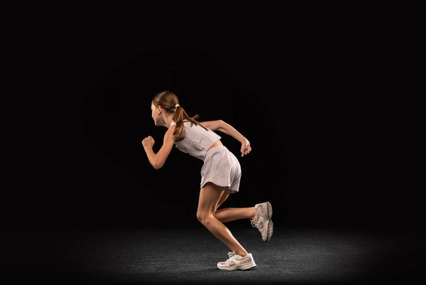 Full-length portrait of girl, teenager running, training isolated over black background. Marathon. Side view - Foto, afbeelding