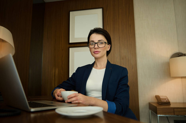 Confident portrait of a successful businessman, prosperous female sales manager looking at camera while sitting at the table and drinking coffee in her office and writing a progress report on laptop - Foto, Bild