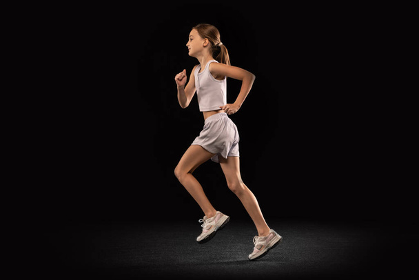 Full-length portrait of girl, teenager, athlete training running isolated over black background. Side view - Foto, afbeelding