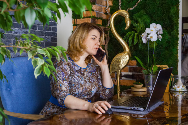 A young woman interior designer is working and talking on the phone in her office at a computer. Green office, live plants in an office space, a wall made of stabilized moss, epoxy resin table - Photo, Image