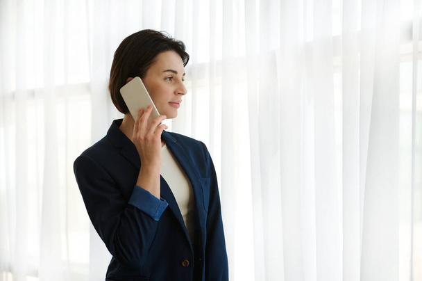 Pewna siebie młoda kobieta ze stylowymi krótkimi włosami w biznesowym garniturze rozmawiająca przez telefon komórkowy stojąc przy oknie w pokoju hotelowym patrząc przez okno na pejzaż miasta podczas podróży służbowej - Zdjęcie, obraz