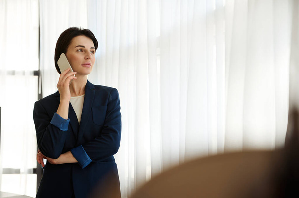 Ευσεβής νεαρή γυναίκα με επαγγελματικό κοστούμι που μιλάει σε ένα κινητό τηλέφωνο, στέκεται δίπλα στο παράθυρο σε ένα δωμάτιο ξενοδοχείου, κοιτάζοντας προσεκτικά από αυτό στην απόσταση στο αστικό τοπίο κατά τη διάρκεια του επαγγελματικού ταξιδιού της - Φωτογραφία, εικόνα