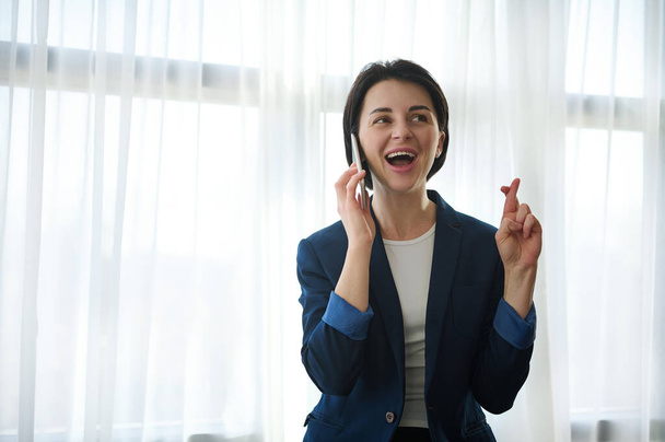 Linda mujer europea alegre joven en traje casual de negocios sosteniendo los dedos cruzados durante la conversación telefónica, sonriendo, mirando a un lado - Foto, imagen