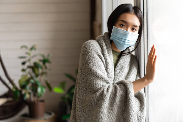 Sick asian girl in medical face mask standing by the window and yearning to go outside, being on quarantine, ill with covid-19 or flu - Photo, Image