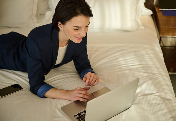 Wysoki kąt widzenia atrakcyjnej pewności siebie odnoszącej sukcesy kobiety biznesu pracującej na laptopie leżącej na łóżku w domu - Zdjęcie, obraz
