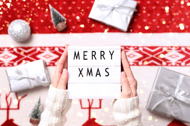 Κοντινό πλάνο του lightbox με κείμενο χαρούμενα Χριστούγεννα στα χέρια και εορταστικό φόντο με χριστουγεννιάτικα δώρα, μινιατούρες δέντρα. Άνω όψη. - Φωτογραφία, εικόνα