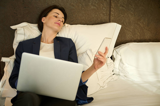 Tired European young business woman using mobile phone and laptop computer while lying down on bed, enjoying relaxation in a hotel room after a business meeting with business partners at work trip - Foto, afbeelding