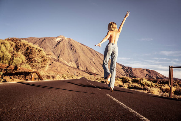 Free happy woman jumping over blue sky and having fun on the empty asphalt road near Teide volcano. Tenerife. Conceptual photo. Real people. Tourist. Traveler. Feminist - Фото, изображение