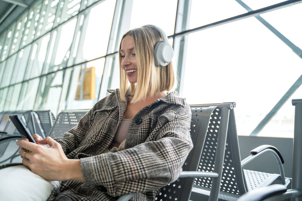 Blanke glimlachende vrouw met smartphone en koptelefoon op de luchthaven terminal zitten en wachten op een vliegtuig, kijken naar iets op sociale media. Reiziger. Toerist. vliegvervoer.  - Foto, afbeelding