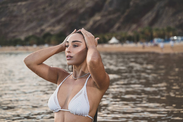Beautiful spanish natural woman in white fashionable bikini posing in the ocean, on the sunset beach. Wet skin and hair. Skin care. Summer island vibes. - Foto, immagini