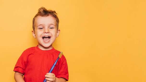 Le garçon heureux tient une brosse à dents bleue sur un fond jaune et sourit en montrant ses dents. Soins de santé, hygiène buccodentaire. Un endroit pour votre texte. - Photo, image