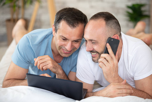 smiling gay couple using laptop and phone on bed - Fotoğraf, Görsel