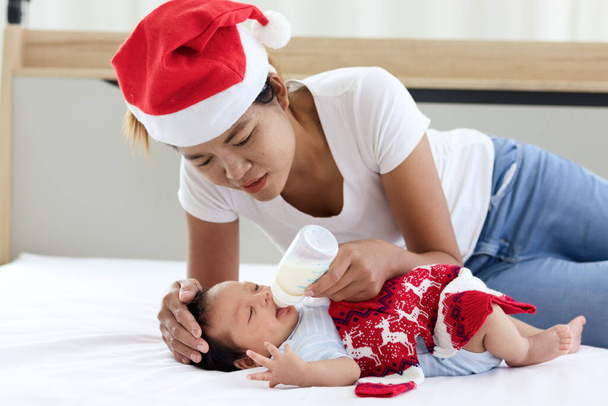 Un pequeño bebé recién nacido adorable dulce niña acostada en la cama blanca con mamá que usa un sombrero rojo de Navidad de Santa y sostiene el biberón de leche para alimentar al bebé, la madre muestra amor y cuidado a un nuevo miembro de la familia - Foto, imagen