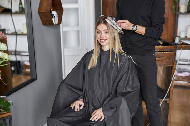 Confident male stylist is dyeing hair of blond caucasian female client - Foto, afbeelding