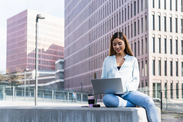 Smiling brunette woman working outdoors surrounded by buildings with laptop and earphones. Business concept - Photo, Image