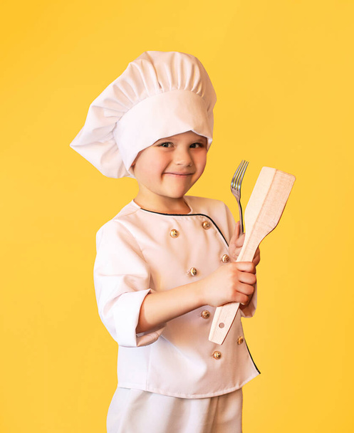 Smiling child in white chef uniform,holding wooden spatula and fork in hands. Cheerful yellow background. - Foto, Imagem