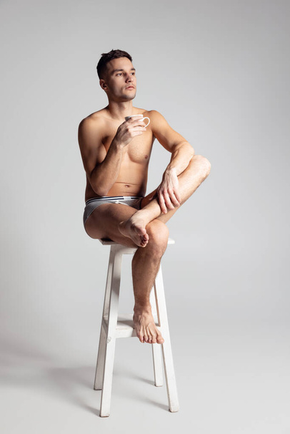 Portrait of young handsome shirtless man sitting on chair isolated on gray studio background. Natural beauty of male body - Foto, imagen