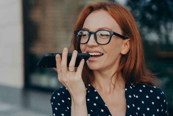 屋外に立っている間に仮想デジタル音声アシスタントを使用して笑顔の赤い髪のビジネス女性 - 写真・画像