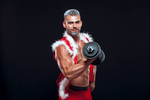 Sexy Santa Claus. Bodybuilder young handsome santa clause smile holds a dumbbells and shows off abs cubes at New Years eve and Christmas winter holiday black background. - Photo, image