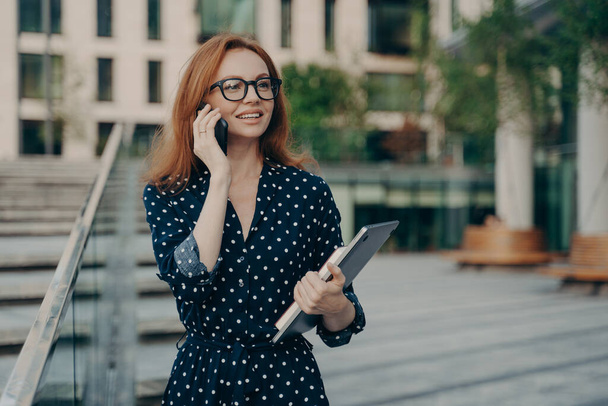 Positive redhead European woman has telephone conversation while walking outdoor - Φωτογραφία, εικόνα