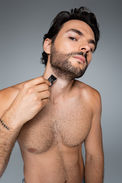 tattooed and shirtless man trimming beard isolated on grey - Foto, Imagem