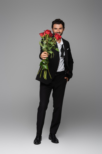 longitud completa de hombre feliz en traje sosteniendo rosas rojas mientras está de pie con la mano en el bolsillo en gris - Foto, Imagen
