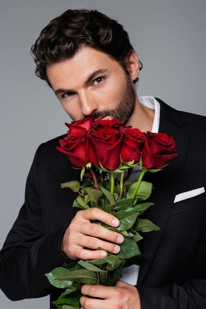 bearded man in formal wear smelling red roses while looking at camera isolated on grey - Photo, Image