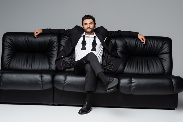 confident man in suit sitting on black leather sofa on grey - Foto, Bild
