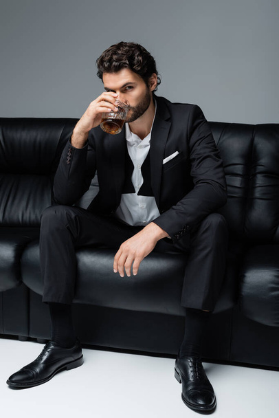young bearded man in formal wear sitting on black sofa and drinking whiskey on grey - Fotoğraf, Görsel