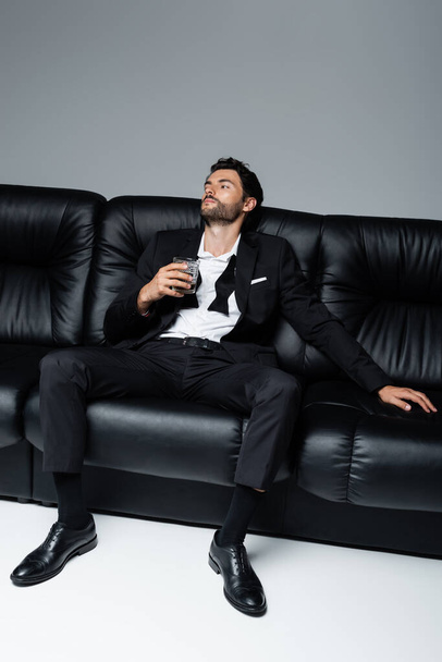 relaxed man in formal wear sitting on black sofa and holding glass of whiskey on grey - Φωτογραφία, εικόνα