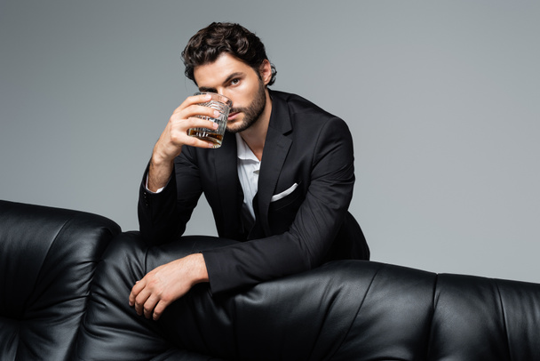 man met baard in pak staat in de buurt van een zwarte bank en houdt glas whisky geïsoleerd op grijs - Foto, afbeelding