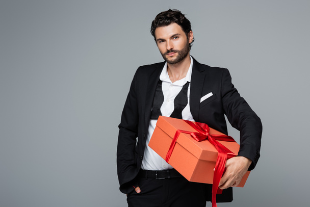 bearded man in suit holding red gift box and standing with hand in pocket isolated on grey  - Photo, Image
