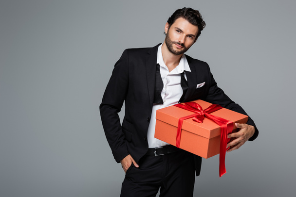 bearded man in suit posing with red gift box and standing with hand in pocket isolated on grey  - Photo, Image